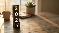 Wooden blocks stacked vertically spelling 2027 on a rustic table with coffee cup and plant in warm morning light.