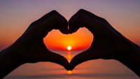 Hands forming heart shape silhouette against vibrant sunset sky with sun visible through fingers creating romantic love symbol for Valentine's Day cards.