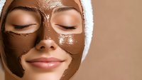 Young woman with chocolate facial mask wearing white towel on head during spa skincare treatment, close up view on relaxed face with closed eyes.