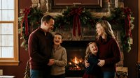 Happy family of four celebrating Christmas together by decorated fireplace with festive garland and red ribbons in cozy living room during holiday season.