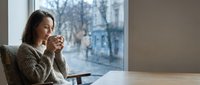 Woman with long dark hair and South Asian appearance sitting at table by window, holding coffee cup, looking outside at winter street scene, contemplative mood.