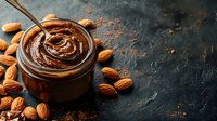 Creamy chocolate almond butter spread in glass jar with spoon swirl and scattered almonds on dark stone background. Food photography for packaging design.