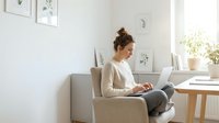 Young Caucasian woman working on laptop in modern minimalist home office with natural light, comfortable armchair and wooden desk setup for productivity.