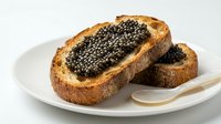 Toasted bread slices topped with creamy spread and black caviar served on white plate with spoon for gourmet appetizer.