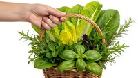 Hand holding wicker basket filled with fresh green herbs including basil, rosemary, and leafy vegetables for cooking and culinary use.