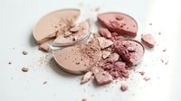 Broken makeup powder compacts in nude and pink shades with crumbled cosmetic powder scattered on white background for beauty and fashion concepts.