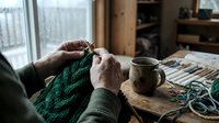 Senior woman knitting green wool sweater at wooden table with coffee cup in cozy home interior during winter afternoon crafting session.