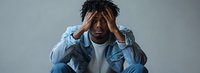 Young African American man in denim jacket showing signs of stress, anxiety, and emotional distress while sitting against gray background, head in hands.