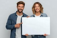 Cheerful young couple in denim shirts pointing at blank white poster against gray background, perfect for adding custom text or promotional content.