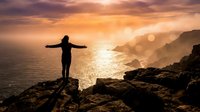 Silhouetted female figure standing on rocky cliff edge with arms outstretched overlooking golden sunset ocean and coastal landscape, conveying freedom, achievement, and contemplation.