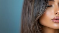 Young Asian woman with glossy straight brown hair and pink lipstick against blue background, closeup beauty portrait showing flawless glowing skin and natural makeup.