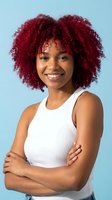 Young African American woman with vibrant red curly hair smiling confidently with arms crossed wearing white tank top against blue background.