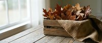 Autumn leaves in rustic wooden crate with burlap fabric on white wooden table near window. Seasonal home decor and fall harvest decoration concept.
