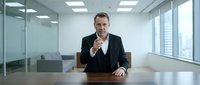 Professional middle-aged Caucasian businessman in black suit sitting at conference table pointing finger at camera in modern glass office boardroom.