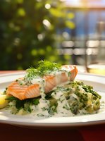 Grilled salmon fillet with creamy dill sauce served on mashed potatoes and fresh herbs, garnished with lemon slice on white plate in restaurant setting.