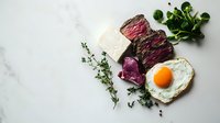 Medium rare sliced beef steak with sunny side up egg, fresh herbs and pickled vegetables on marble background. Protein-rich breakfast concept.