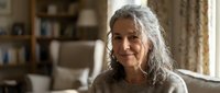Smiling mature woman with gray curly hair sitting comfortably in cozy living room interior, expressing contentment and wisdom in warm home environment.