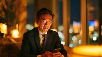 Middle-aged Asian businessman in suit using smartphone at night in urban city street with warm bokeh lights for corporate technology and communication concepts.