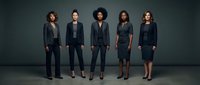 Diverse group of five professional men and women wearing formal black business attire standing together against neutral gray background for corporate team, leadership, and workforce concepts.