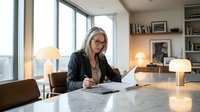 Mature businesswoman with gray hair reviewing documents at modern office desk with warm lighting and city view windows for corporate presentations.