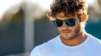 Young Caucasian man with curly dark hair and stubble wearing sunglasses and white t-shirt smiling outdoors in natural sunlight against blurred background.