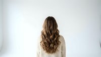 Young woman with long wavy brown hair wearing beige knit sweater standing with back turned against white background for lifestyle design.