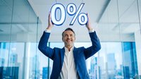 Male businessman in formal blue suit and white shirt holding zero percent symbol above head in modern office with city skyline view, representing zero interest rates, no fees, or free promotional offers for financial services and business concepts.