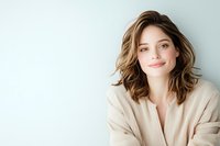 Young Caucasian woman with wavy brown hair and natural makeup wearing beige cardigan smiling warmly against light blue wall background. Wellness and natural beauty concept.
