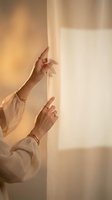 Woman with medium skin tone wearing beige linen shirt and gold bracelets, gently touching sheer white curtain with both hands in warm golden sunlight, conveying calm and serenity.
