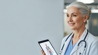 Senior female doctor with gray hair using tablet for telemedicine consultation in modern medical office, smiling confidently while providing healthcare.