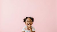 Young child with dark curly hair and medium skin tone, wearing white shirt, smiling with hand near mouth against soft pink background.