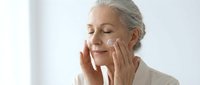 Senior Caucasian woman applying anti-aging cream to face with gentle massage motions in bright bathroom for mature skincare routine and wellness.