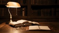 Vintage banker lamp illuminates open books and reading glasses on wooden desk in classic library study room with bookshelf background for academic research.