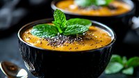 Creamy pumpkin soup garnished with fresh mint leaves and black chia seeds served in black ceramic bowl, close up view on dark background. Food photography.