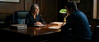 Female lawyer or judge in professional attire conducting serious meeting with male client across wooden desk in law office with documents and desk lamp.
