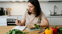 Young Caucasian woman eating healthy salad bowl while working on laptop in modern kitchen, balanced lifestyle and nutrition concept for busy professionals.