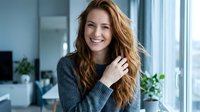 Young attractive Caucasian woman with long wavy brown hair smiling warmly in modern office space with natural lighting and plants in background.
