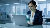 Professional woman with dark hair and olive complexion wearing navy blazer works at laptop in modern glass office with holographic security interface overlay, representing cybersecurity, data protection, and corporate technology solutions.