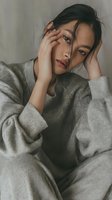 Young Asian woman experiencing headache or stress holding head in hands wearing cozy gray sweater in soft natural lighting for wellness and mental health concepts.