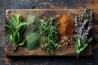 Fresh culinary herbs and colorful spice powders arranged on rustic wooden cutting board, including sage leaves, spirulina powder, oregano, paprika and lavender for cooking and garnishing.
