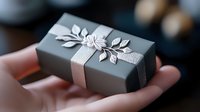Elegant gray gift box decorated with silver ribbon and delicate metallic leaves held in palm, perfect for special occasions and luxury presentations.
