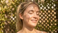 Young woman with light skin tone and blonde hair smiling outdoors with dappled sunlight on her face, suitable for wellness, beauty, and lifestyle marketing campaigns.