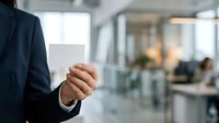 Businessman in dark suit holding blank white business card in modern office environment with blurred colleagues working in background.