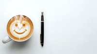 Coffee cup with smiley face latte art and black pen on white background. Morning motivation workspace setup for productivity and positive mindset.