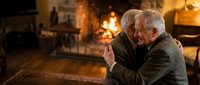 Elderly Caucasian man in warm sweater sitting by cozy fireplace, enjoying hot beverage in comfortable living room during winter evening at home.