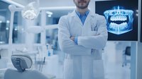 Male dentist in white coat standing confidently in modern dental office with digital jaw scan displayed on monitor in background.