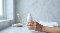 Mature woman with light skin tone holding white cosmetic bottle in bathroom setting with marble countertop, displaying skincare or beauty product for advertising and promotional use.