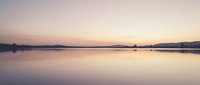 Serene lake at dawn with mirror-like water reflecting soft pink and purple sky, distant treeline silhouettes, calm nature landscape for meditation and relaxation projects.