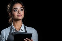Young Hispanic female restaurant worker in white shirt and dark apron holding digital tablet, looking confident against black background. Professional service industry portrait.