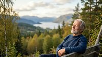 Senior man with gray hair and light complexion, wearing dark blue sweater, relaxing outdoors on wooden bench overlooking mountain lake surrounded by evergreen forest and birch trees during golden hour.